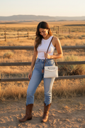 Woman in western outfit with cowboy boots carrying white cowhide RODEO crossbody bag at golden hour ranch setting